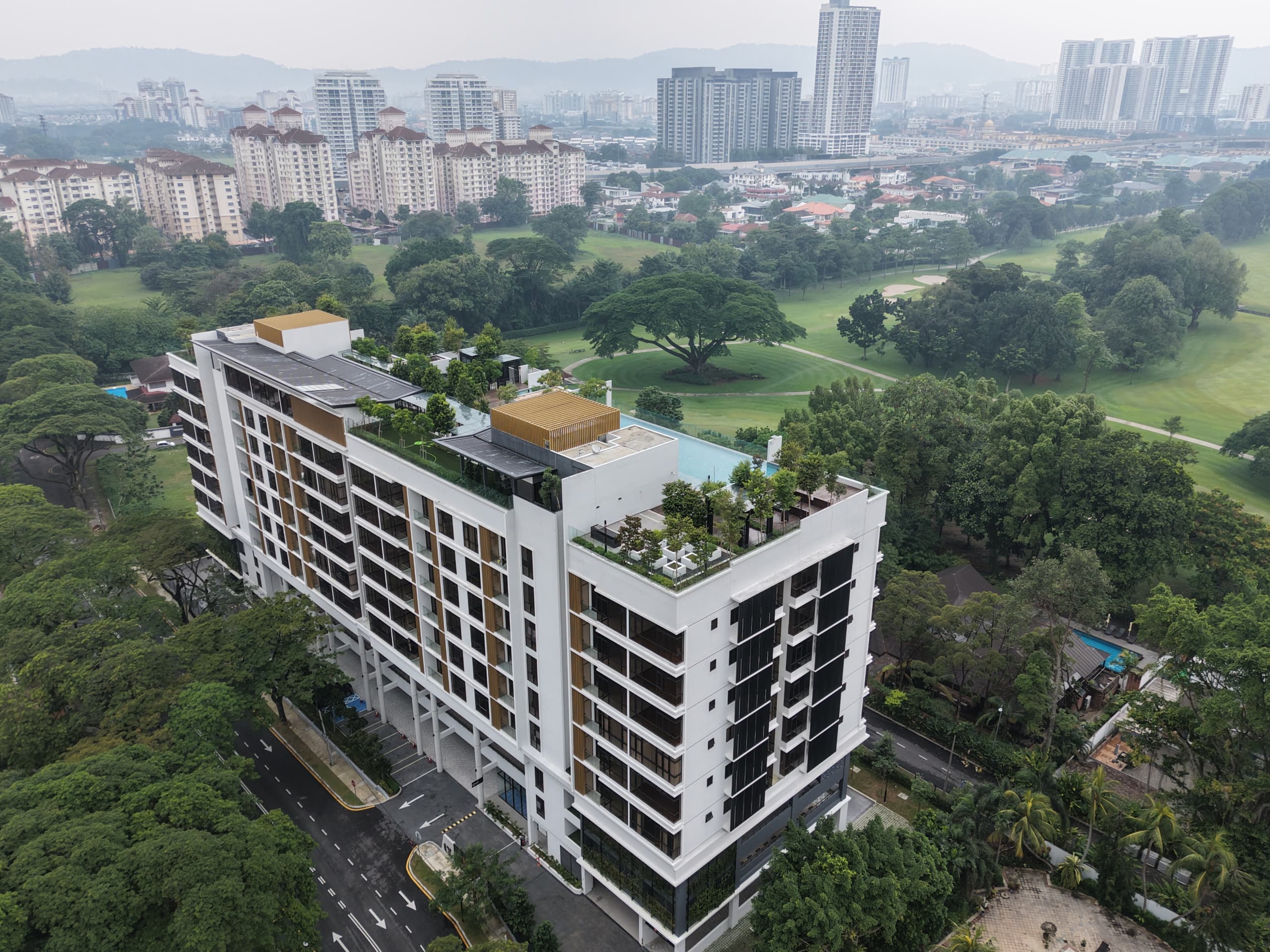 Aerial view of a white mid-rise building with rooftop greenery beside a tree-filled golf course.